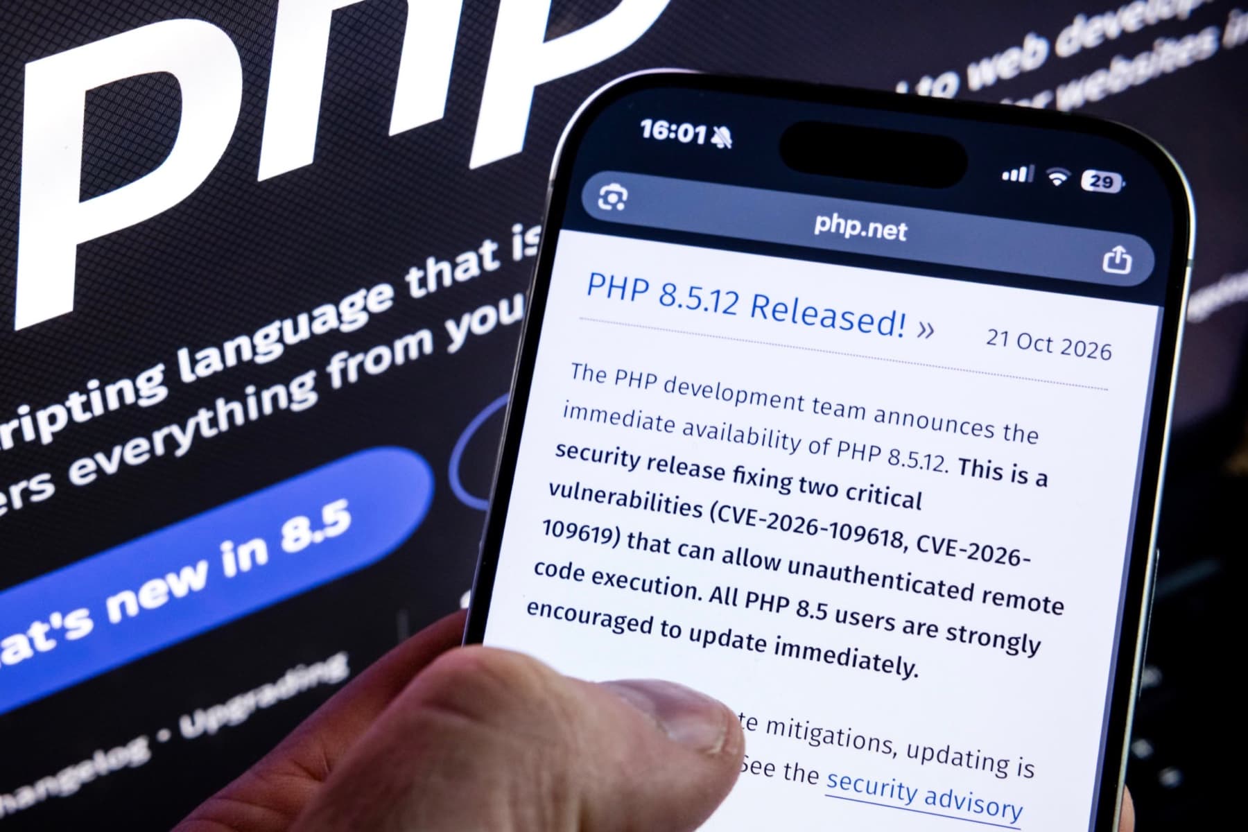 A hand holding a phone showing the php.net page "PHP 8.5.12 Released!", dated 21 October 2026, in front of a large PHP logo on a screen.