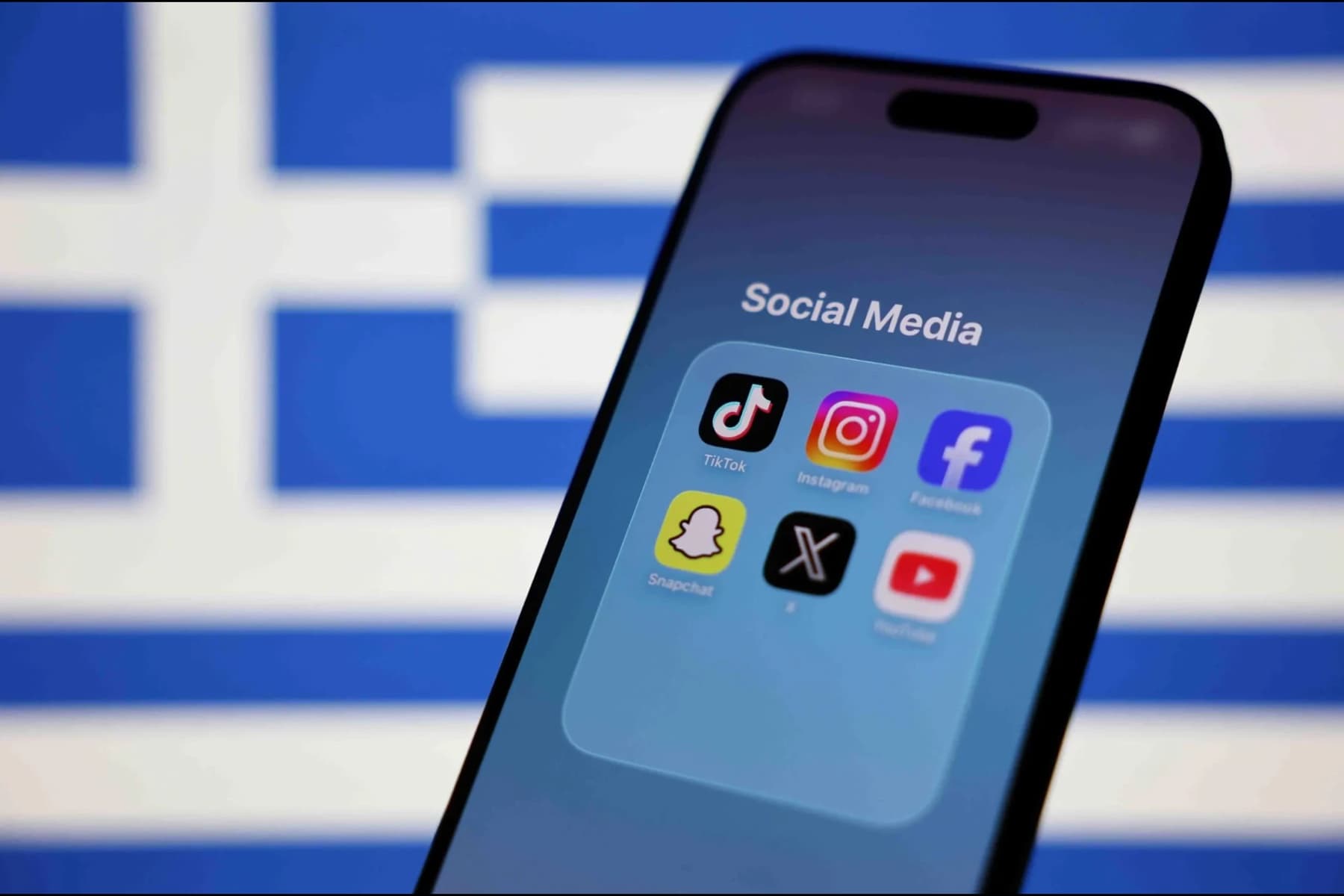A phone showing a "Social Media" folder of app icons, including TikTok, Instagram and Facebook, in front of a blurred Greek flag.