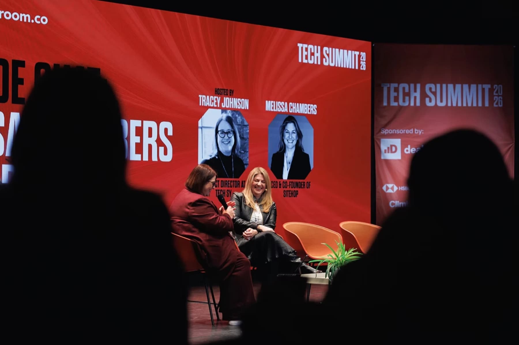 Two women in conversation on a stage in front of a large red screen reading Tech Summit 2026, with audience members silhouetted in the foreground.