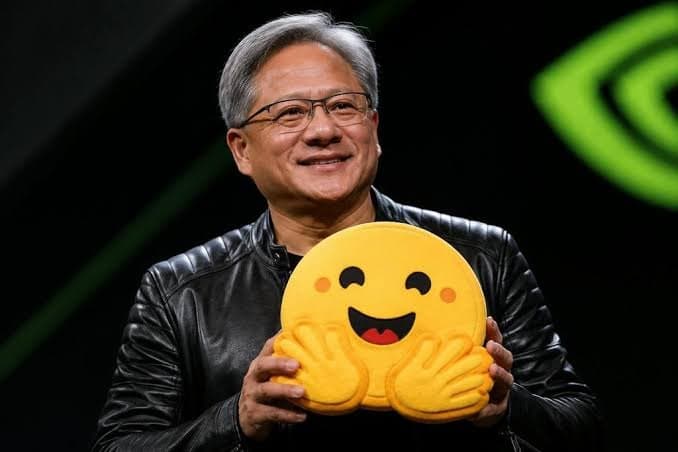Jensen Huang holding a large Hugging Face emoji cushion on stage.