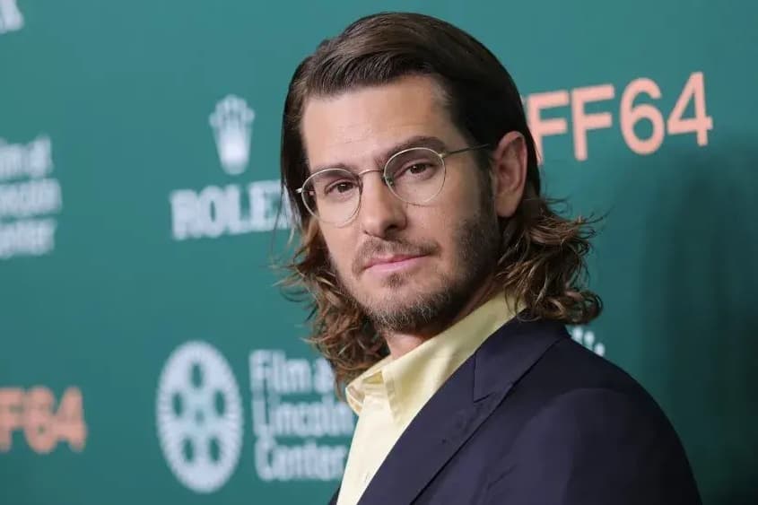 Andrew Garfield, in glasses and a dark suit, on the red carpet at the New York Film Festival in front of a green NYFF64 backdrop.