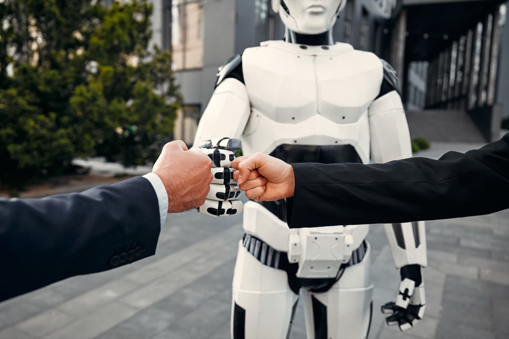 Two people in business suits fist-bumping a white humanoid robot outside an office building.
