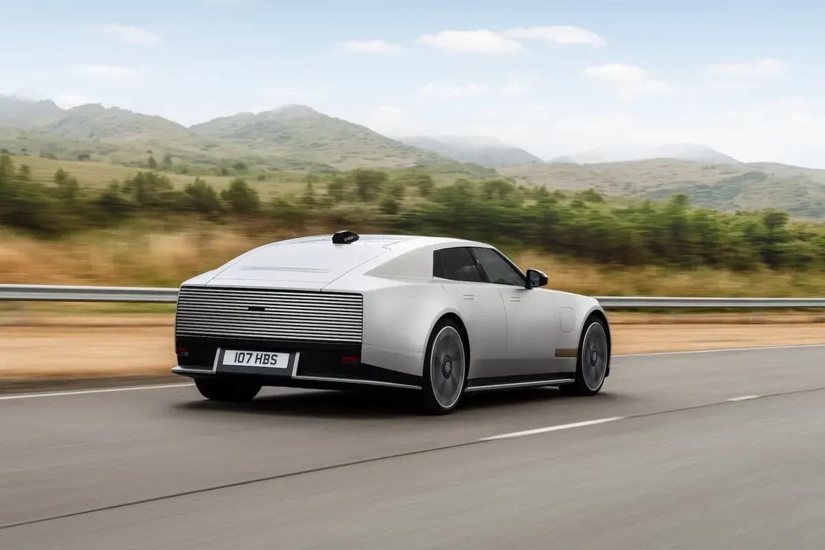 A silver Jaguar Type 01 seen from the rear three-quarter, driving along a road with green hills behind.