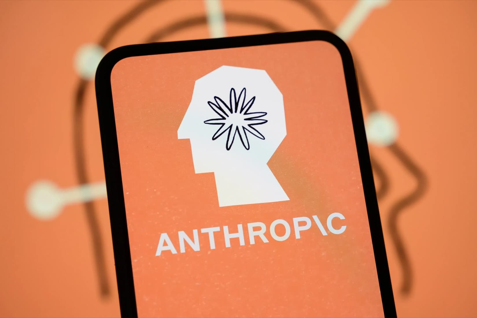 The Anthropic logo on a smartphone screen, held in front of an orange Anthropic graphic.